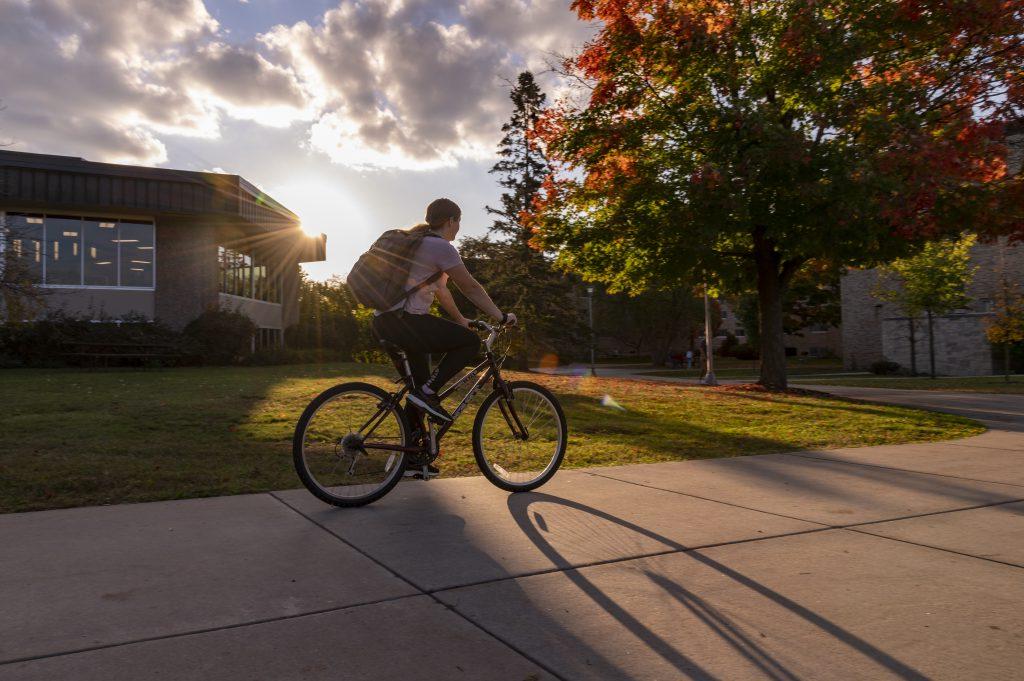 Student riding a bike on the sidewalk by the DeBot Dining Center at sunset.